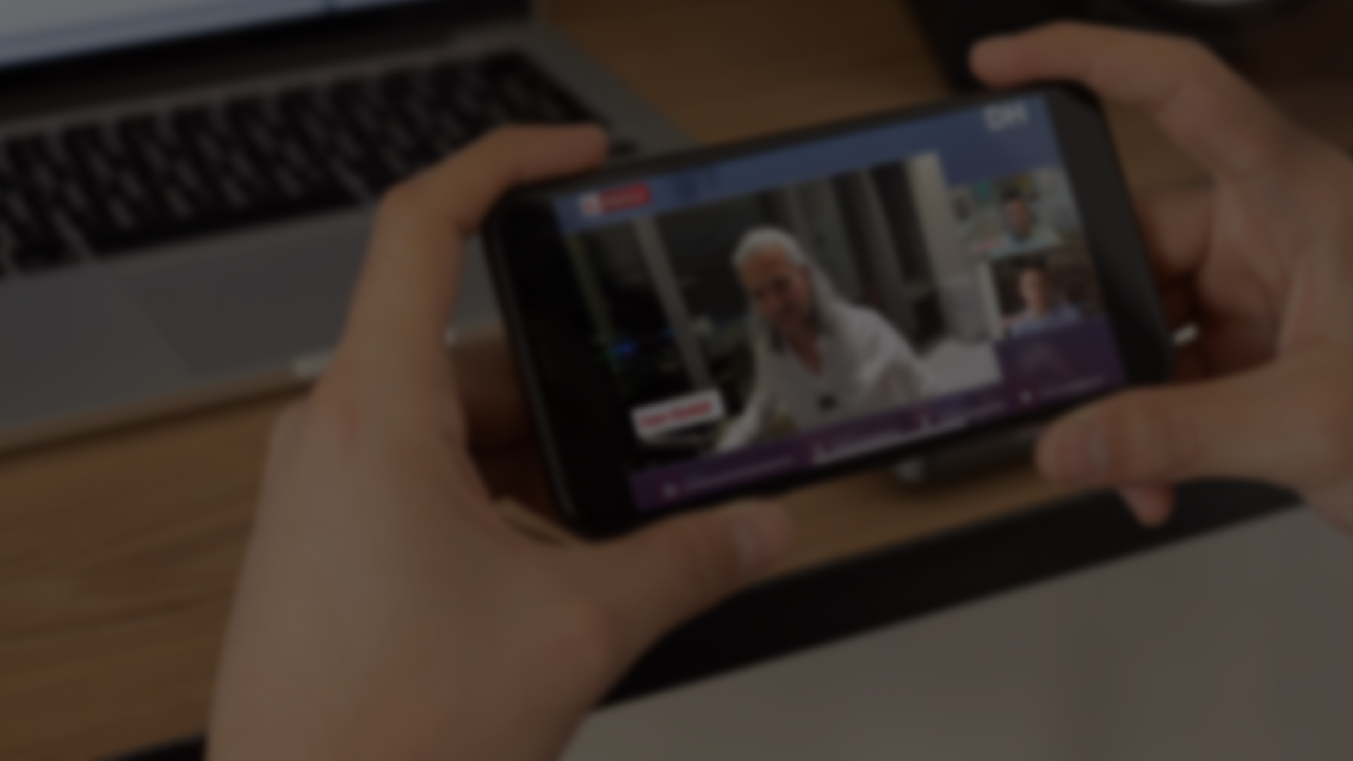 A person is watching a live stream on their smartphone. On the screen, a speaker in a white shirt is visible, along with other participants in smaller windows.